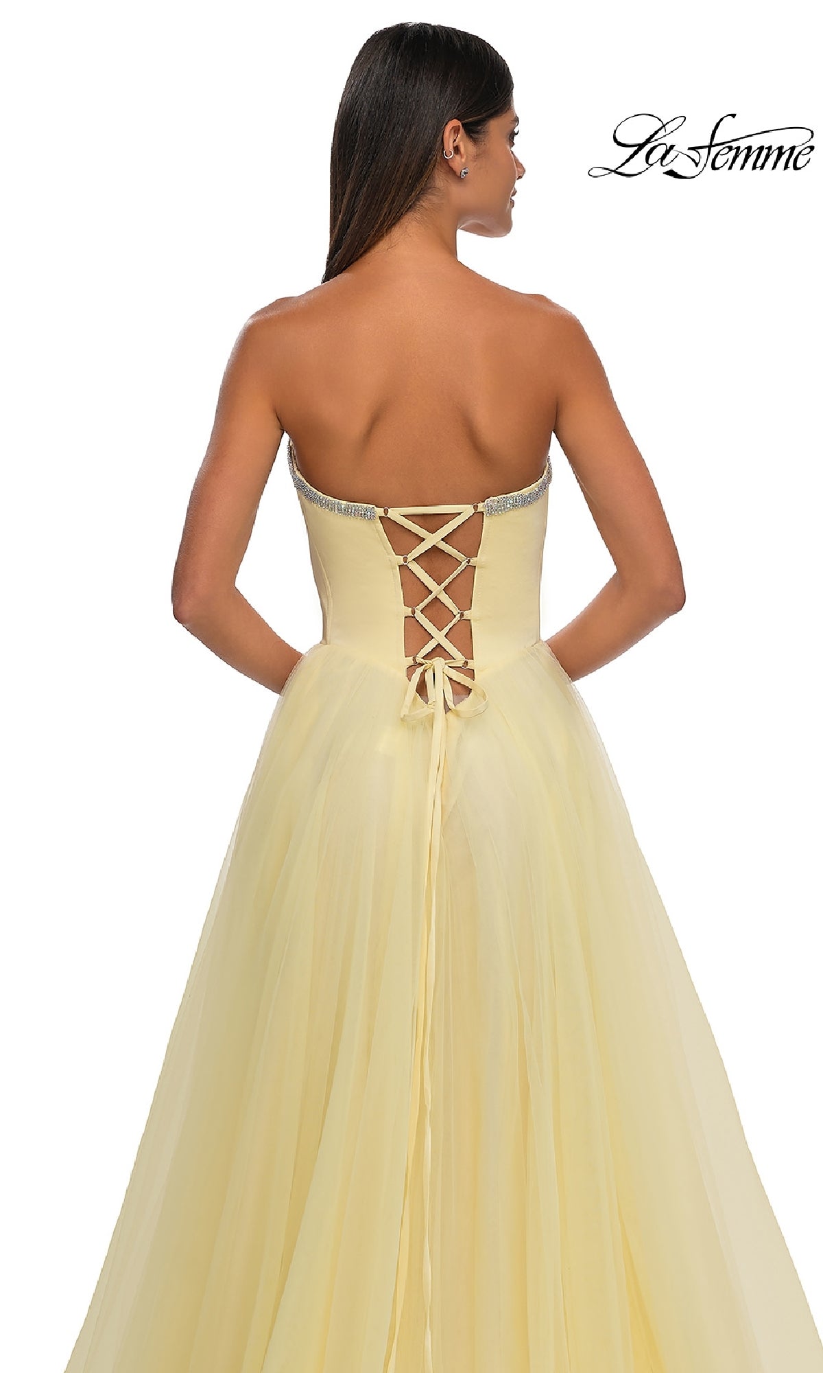 A woman in the pale yellow La Femme 32731 strapless long prom ball gown with a sweetheart neckline and lace-up back stands facing away, showcasing the floor-length, flowing fabric.